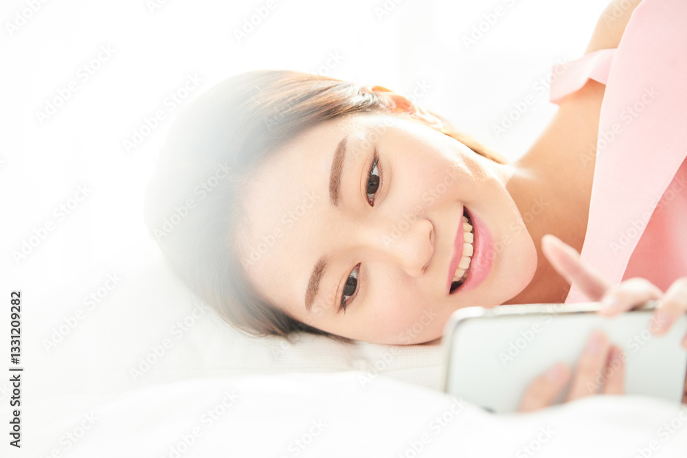 Obraz premium A young and beautiful woman, dressed in pink, is lying on a bed in a sunlit room, smiling happily while looking at her smartphone