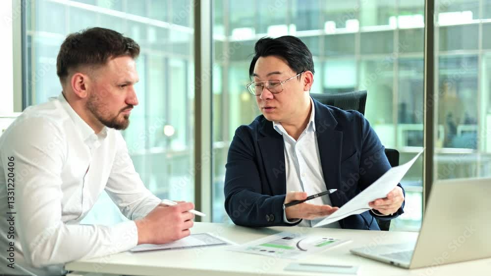Two business men having a discussion in a modern office. One man explains some documents to the other. Natural light comes in through large windows. Two business men having a discussion in a modern office. One man explains some documents to the other. Natural light comes in through large windows.