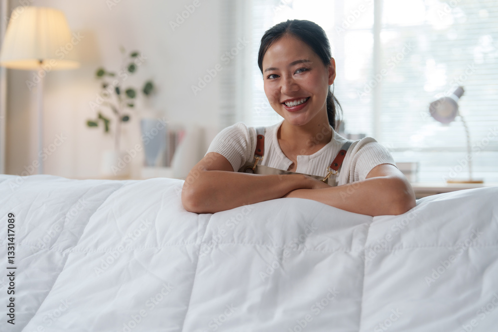 Young, happy housekeeper smiling and leaning against a soft white comforter, enjoying a moment of satisfaction after completing the cleaning of a tidy hotel bedroom Young, happy housekeeper smiling and leaning against a soft white comforter, enjoying a moment of satisfaction after completing the cleaning of a tidy hotel bedroom