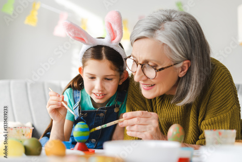 Canvas Print Little happy girl and senior gray haired woman painting easter eggs together at
