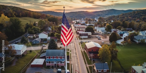 Fototapeta Naklejka Na Ścianę i Meble -  Aerial view of a small American town with the United States flag in the foreground, surrounded by autumn foliage, rolling hills, and a serene sunset in the background