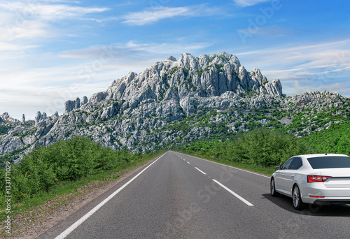 A white car drives along a scenic highway surrounded by lush greenery, heading towards towering rocky mountains under a bright blue sky, capturing the essence of adventure and exploration.