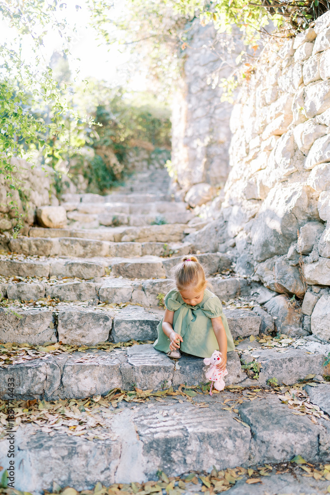Fototapeta premium Little girl squats on old stone steps in the garden playing with a toy mouse