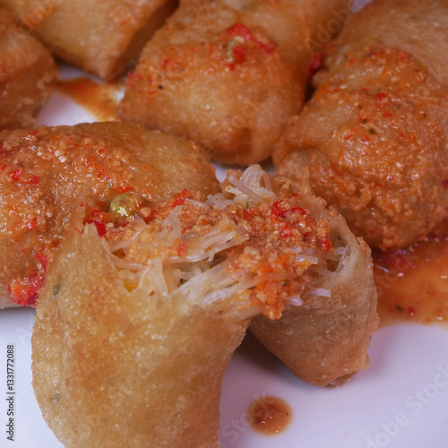 Close-up golden crispy risol or risoles kampung filled with vermicelli, coated in spicy chili sauce, served on a white plate. A savory and spicy Indonesian delicacy.