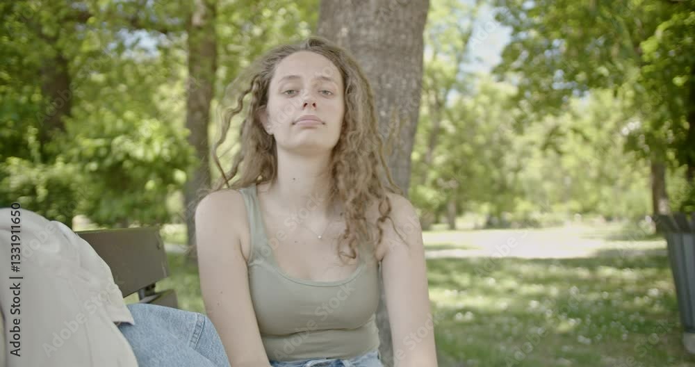 Young woman sitting on a park bench enjoying the summer weather. She brushes a strand of hair out of her face as she takes a deep breath and looks into the distance. Young woman sitting on a park bench enjoying the summer weather. She brushes a strand of hair out of her face as she takes a deep breath and looks into the distance.