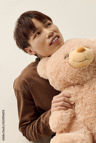 Young handsome Asian man joyfully holds a soft teddy bear in a cozy indoor setting