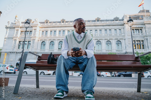 Obraz na płótnie fashionable Black male sits on bench in urban setting, holding smartphone and glancing to the side. His relaxed yet stylish outfit, highlights modern street fashion and digital connectivity. w salonie