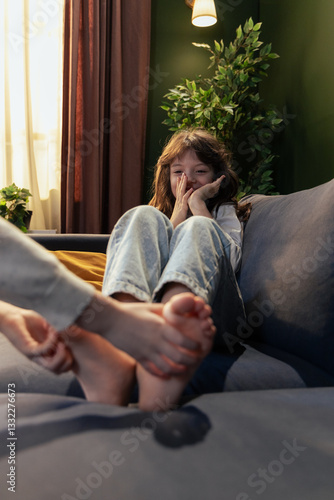 Fotografie Mother massaging daughter's feet on sofa, girl giggling and hiding face