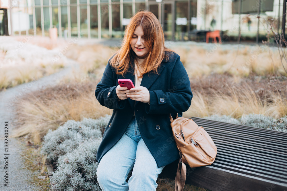 Happy woman sitting on the bench and chatting on phone outdoors. Urban, people concept. Smiling 30s redhead woman text messaging on smart phone at sunny day on the city street, spring time