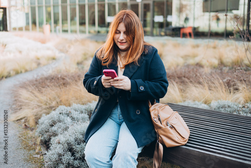 Obraz na płótnie Happy woman sitting on the bench and chatting on phone outdoors. Urban, people concept. Smiling 30s redhead woman text messaging on smart phone at sunny day on the city street, spring time w salonie