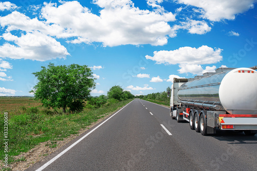 Obraz na płótnie A large fuel tanker truck travels along a straight, open highway surrounded by lush greenery and a bright blue sky with scattered white clouds, reflecting the peaceful countryside landscape. w salonie
