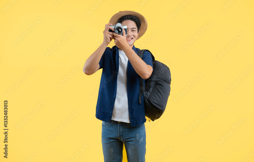 Portrait of young asian man wearing backpack and using camera posing on yellow background Portrait of young asian man wearing backpack and using camera posing on yellow background