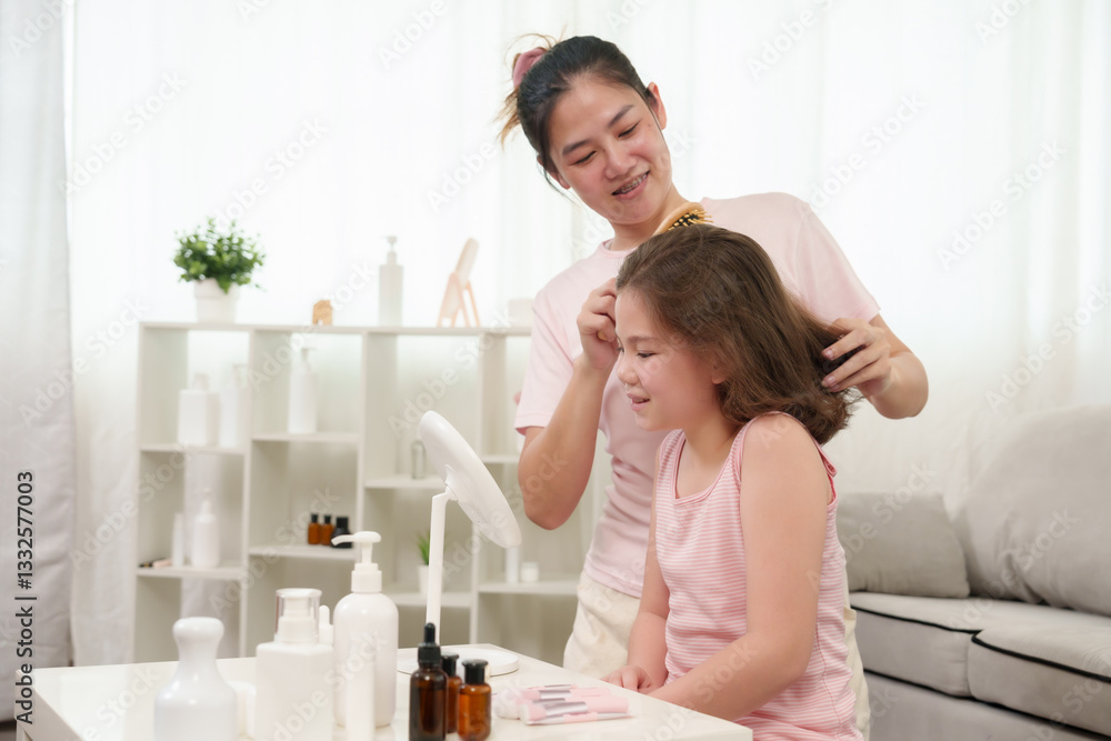 Asian mother brushing Caucasian daughter's wavy brown hair while sitting together at white table. Beauty products, skincare items, and hair accessories arranged neatly in cozy home setting. Asian mother brushing Caucasian daughter's wavy brown hair while sitting together at white table. Beauty products, skincare items, and hair accessories arranged neatly in cozy home setting.