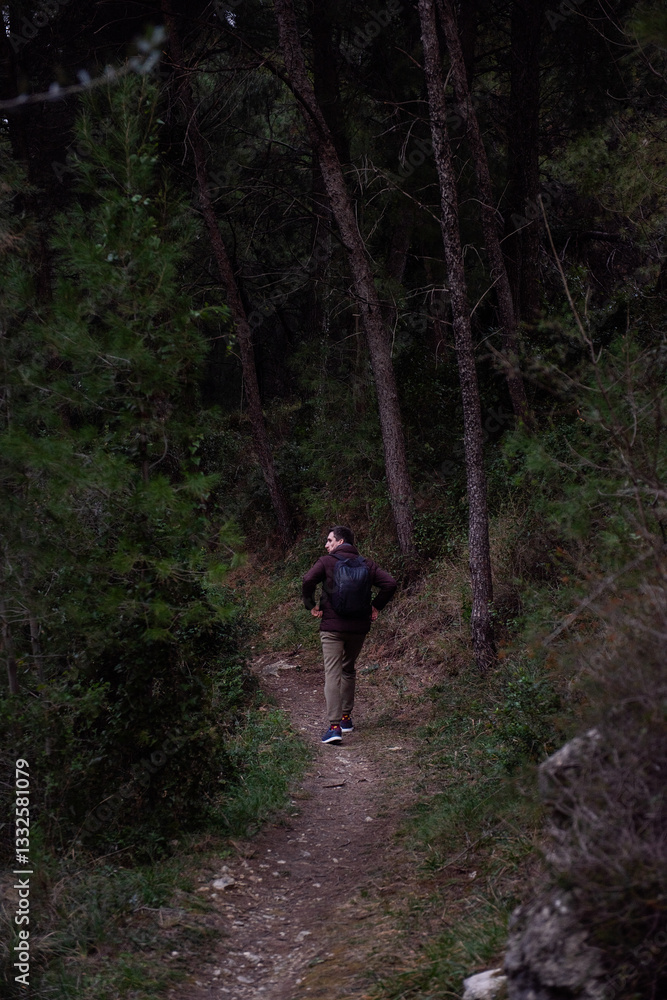 Man wearing a brown jacket and backpack stands on a narrow dirt trail in a dense forest, surrounded by tall trees and lush greenery, creating a peaceful outdoor scene. Man wearing a brown jacket and backpack stands on a narrow dirt trail in a dense forest, surrounded by tall trees and lush greenery, creating a peaceful outdoor scene.