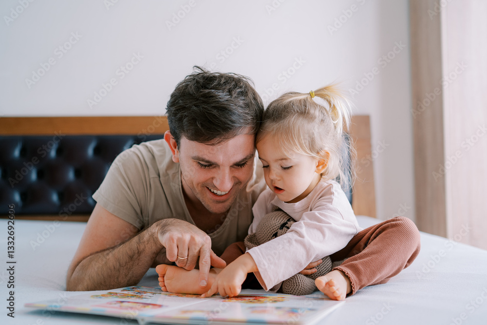 Fototapeta premium Dad and little girl are reading a book of fairy tales on the bed, running their fingers through the pages