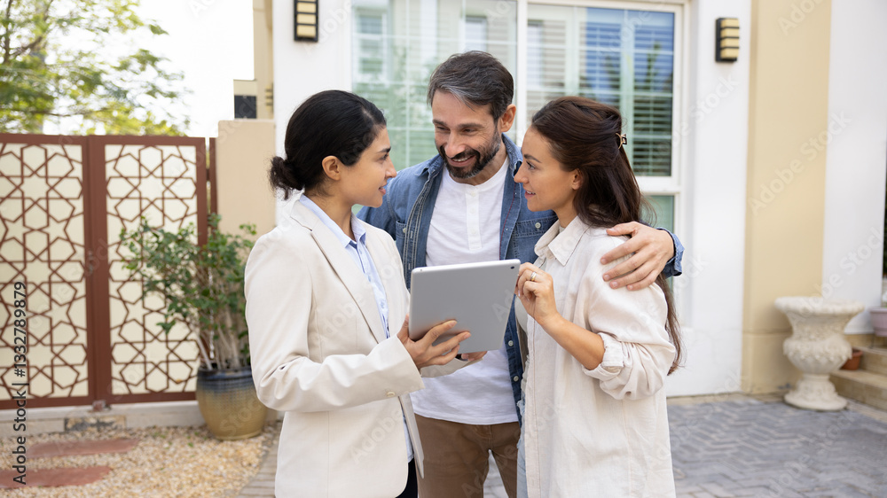 Happy couple meeting with realtor woman at accommodation, standing outdoors on front yard, using digital tablet computer, discussing mortgage terms, house for rent, talking, smiling