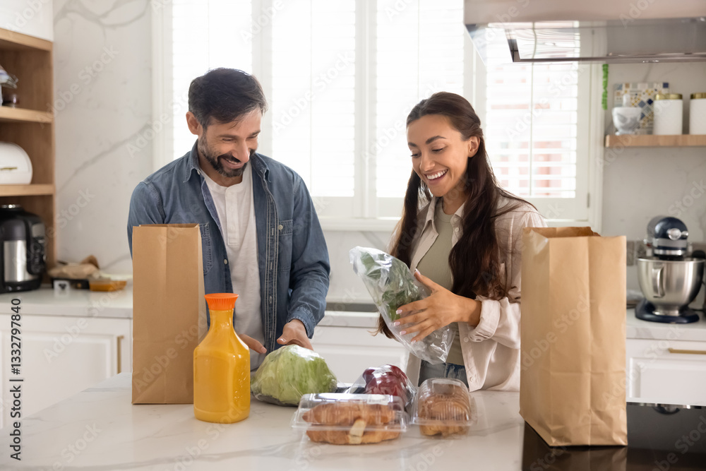 Fototapeta premium Cheerful married couple in love unpacking bags with food from grocery store, talking, laughing, enjoying leisure, weekend at home, domestic activity, producing fresh vegetables from paper packets