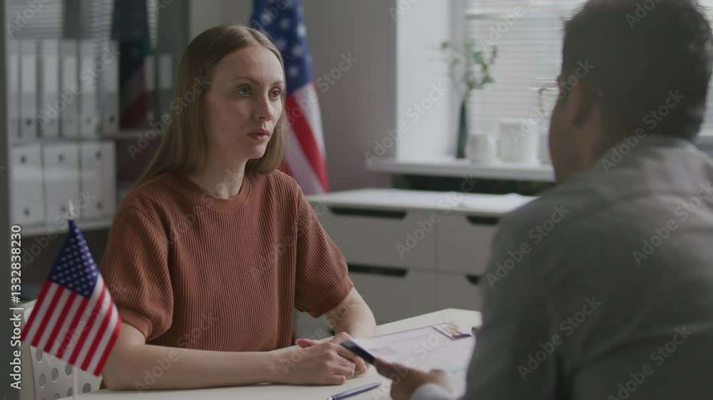 Woman being interviewed by US consular officer reading her application form during visa appointment in government office with American flags Woman being interviewed by US consular officer reading her application form during visa appointment in government office with American flags