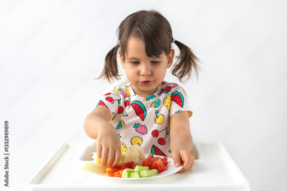 Little girl with pigtails sitting in a high chair, carefully picking up vegetables from a plate. A colorful outfit with fruit prints adds a playful touch to the healthy eating moment. healthy food Little girl with pigtails sitting in a high chair, carefully picking up vegetables from a plate. A colorful outfit with fruit prints adds a playful touch to the healthy eating moment. healthy food