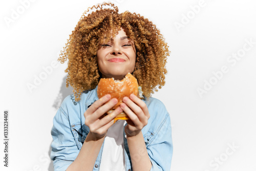 Canvas Print Young woman with curly hair joyfully enjoying a delicious burger against a brigh