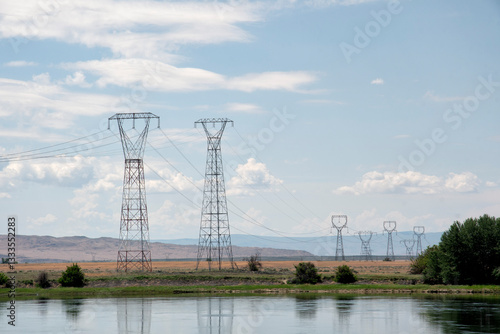 A scenic view showcasing tall power lines stretching across a tranquil landscape, with a river in the foreground and rolling hills under a bright sky.