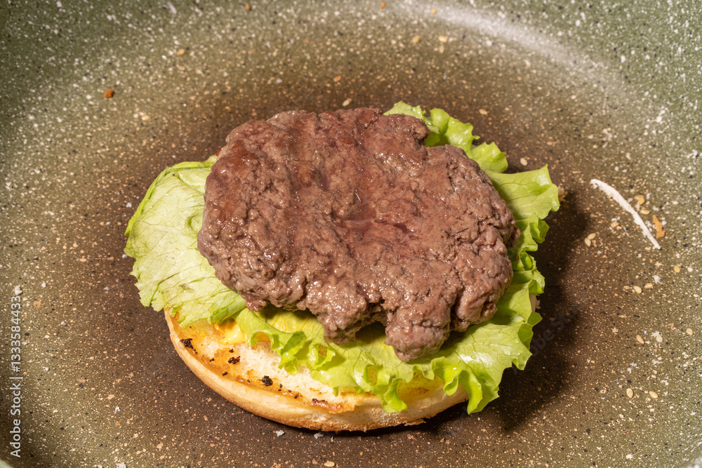 A cooked hamburger patty atop fresh lettuce on a toasted bun in a frying pan, capturing a rustic stage of homemade burger preparation.