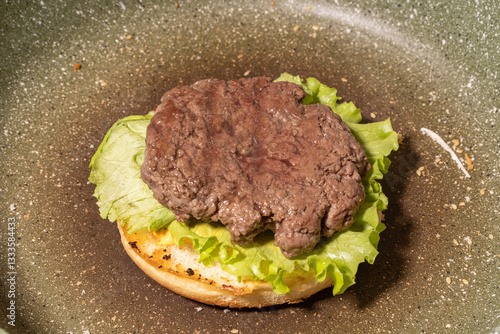 Obraz na płótnie A cooked hamburger patty atop fresh lettuce on a toasted bun in a frying pan, capturing a rustic stage of homemade burger preparation. w salonie