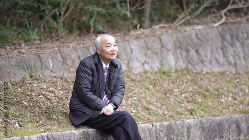 A Japanese winter daytime park. A 78-year-old Japanese man in a suit and a down jacket is sitting on concrete. My son, in his 30s, walks around with a book.