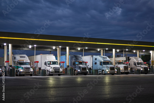 Different big rig semi trucks with turned on headlights and loaded semi trailer standing on the truck stop gas station at night time fill up the trucks tanks to continue their routes in the morning