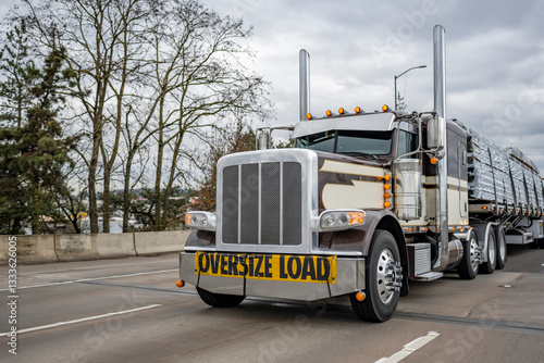 Powerful black low cab profile classic big rig semi truck with oversize load sign on the front transporting fastened cargo on flat bed semi trailer running on the straight wide highway road