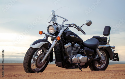 A lone motorcycle stands on the sandy surface of the Namib Desert against a dramatic cloudy sky. The sunset lighting highlights the contrast between nature and machine.