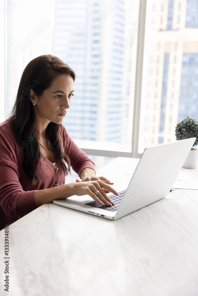 Serious Hispanic businesswoman working at workplace table at office window with urban view, typing on laptop. Employee using modern web technology, wireless online connection for job Serious Hispanic businesswoman working at workplace table at office window with urban view, typing on laptop. Employee using modern web technology, wireless online connection for job