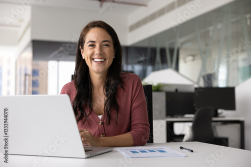 Happy attractive Hispanic business professional woman posing at office workplace alone, working on Internet project, sitting at laptop computer, looking at camera, smiling, laughing