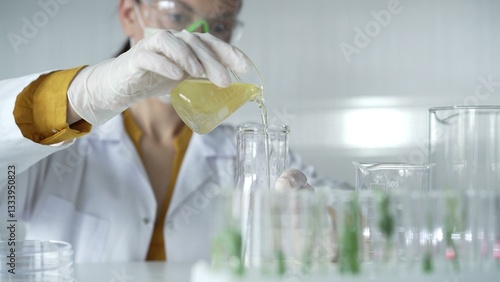 Obraz na płótnie Woman scientist wearing a lab coat, white gloves, and a mask is pouring a yellow oily liquid from a beaker into an Erlenmeyer flask in a laboratory setting, close up. Science and medicine w salonie