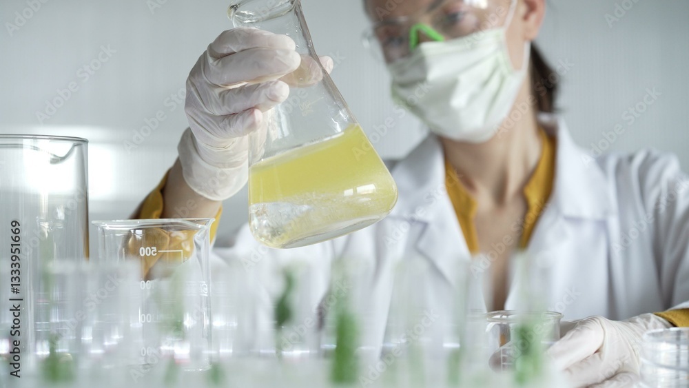 Woman scientist wearing white protective gloves is shaking up a yellow chemical solution inside an Erlenmeyer flask in a laboratory setting, portrait. Science and medicine concepts