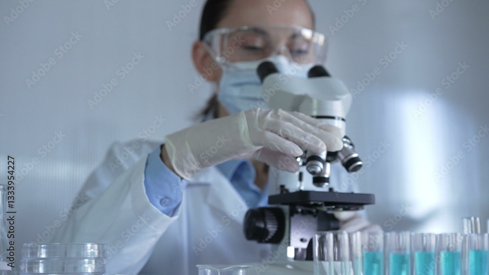 A scientist woman, wearing a lab coat, mask, white gloves and safety glasses, is adjusting a microscope surrounded by test tubes in laboratory A scientist woman, wearing a lab coat, mask, white gloves and safety glasses, is adjusting a microscope surrounded by test tubes in laboratory