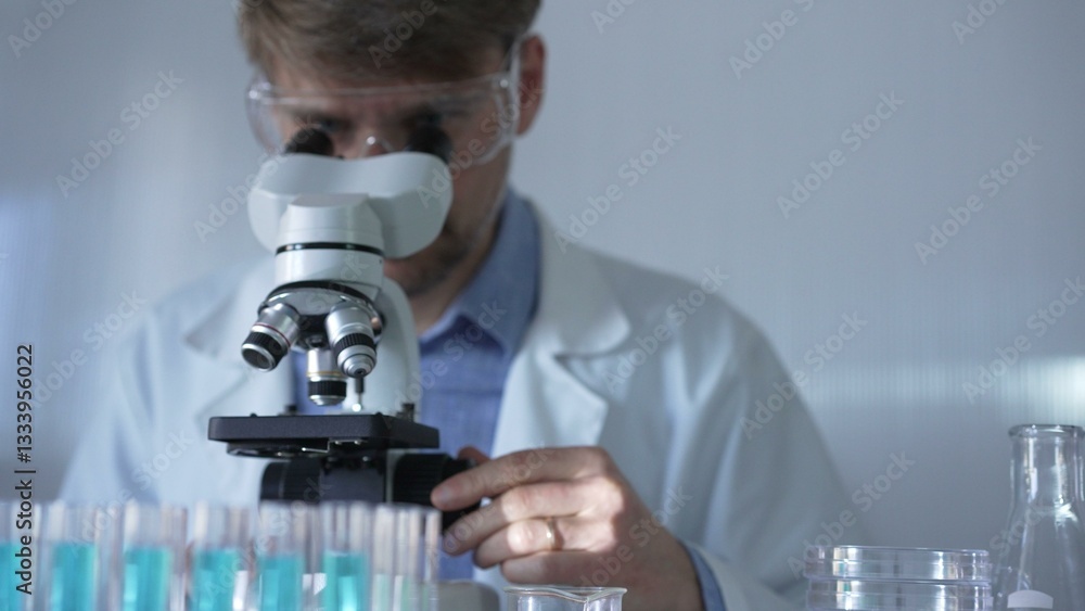 A man scientist, wearing a lab coat, mask, and safety glasses, is using a microscope surrounded by test tubes in laboratory setting. Medicine and science concept