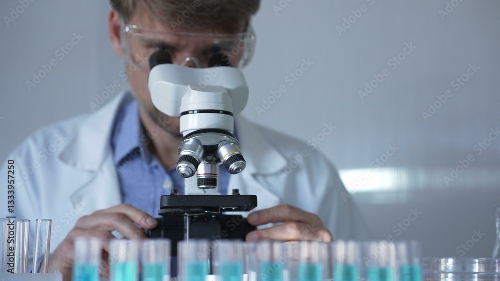 A man scientist, wearing a lab coat, mask, and safety glasses, is using a microscope surrounded by test tubes in laboratory setting. Medicine and science concept