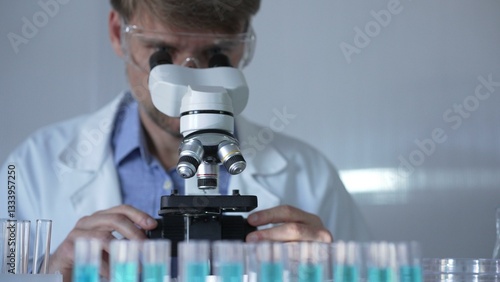 Obraz na płótnie A man scientist, wearing a lab coat, mask, and safety glasses, is using a microscope surrounded by test tubes in laboratory setting. Medicine and science concept w salonie