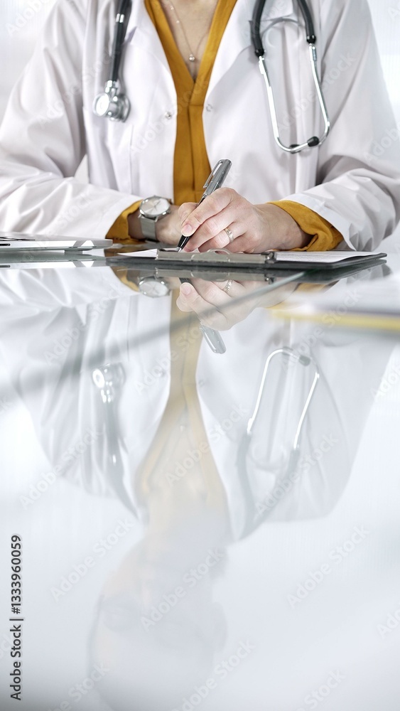 Unknown doctor woman, wearing a stethoscope and yellow blouse under a lab coat, is using a clipboard and taking notes on glass desk in a medical office, close-up vertical view. Medicine concept