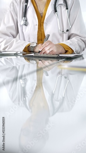 Obraz na płótnie Unknown doctor woman, wearing a stethoscope and yellow blouse under a lab coat, is using a clipboard and taking notes on glass desk in a medical office, close-up vertical view. Medicine concept w salonie