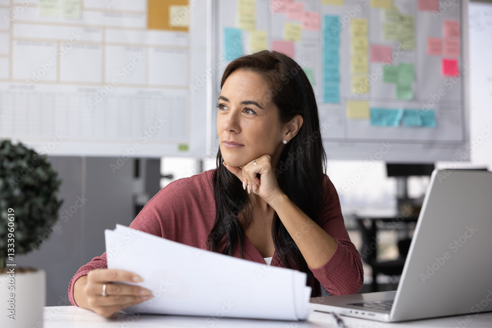 Thoughtful business woman analyzing financial report, sitting at office workplace with laptop, touching chin, looking away, holding paper document, thinking on successful strategy, business plan Thoughtful business woman analyzing financial report, sitting at office workplace with laptop, touching chin, looking away, holding paper document, thinking on successful strategy, business plan