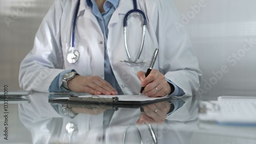 Woman doctor, wearing a blue blouse under a white medical coat, is using a clipboard and taking notes on glass desk in a medical office, close-up. Medicine