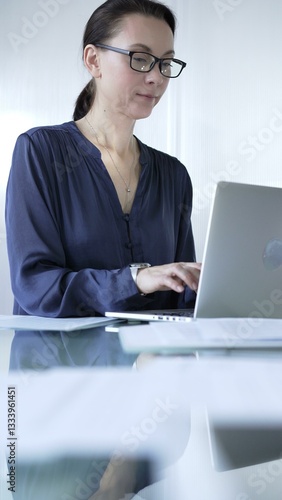 Focused businesswoman with glasses typing on laptop at modern glass desk in well lit office. Business people concept