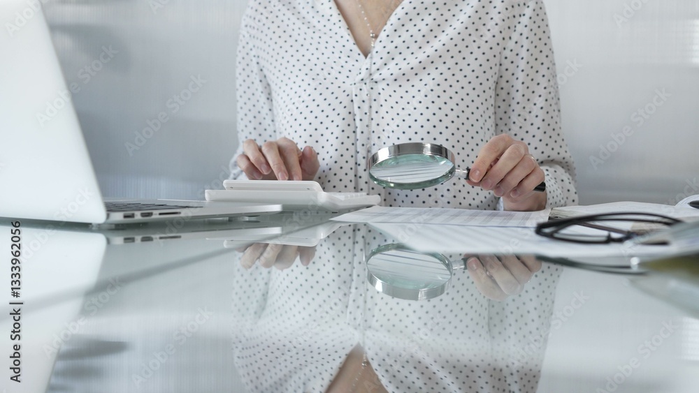 Businesswoman carefully reviewing financial reports with magnifying glass and calculator, conducting audit and analyzing data, closeup view. Taxes and audit in business