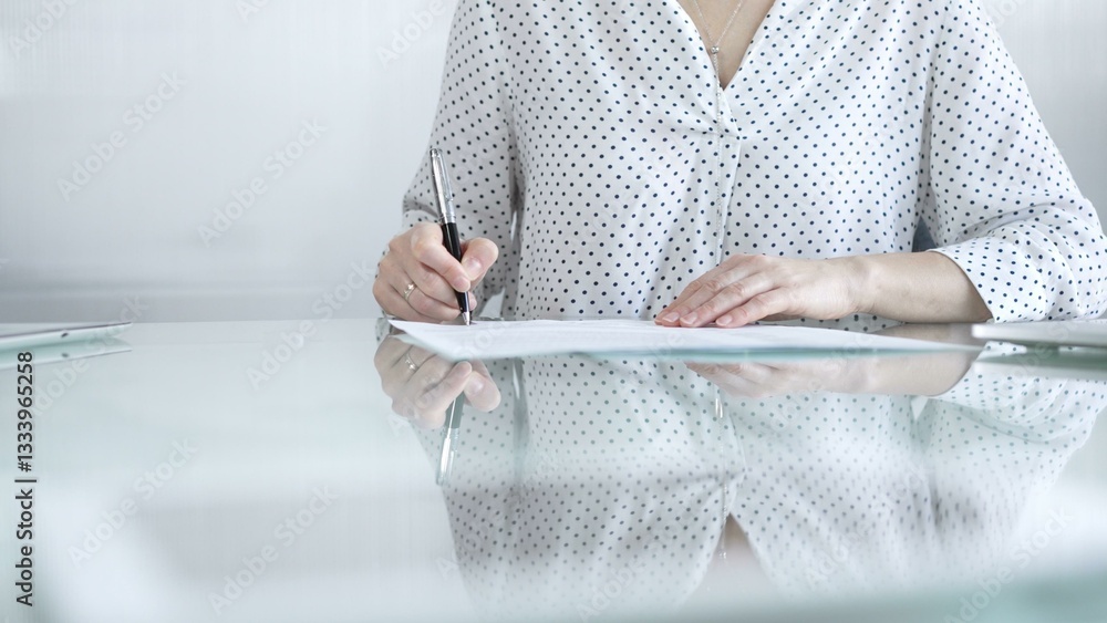 Businesswoman in white dotted blouse is signing a contract at a sleek glass desk in a modern office, showcasing professionalism and focus in her work environment. Business people concept