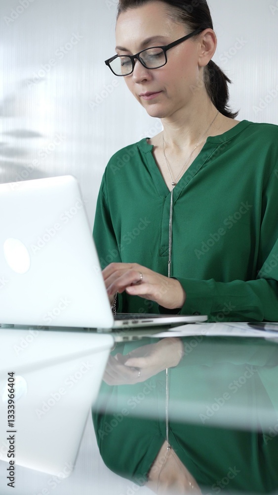 Businesswoman, wearing green blouse, is typing on laptop keyboard, preparing taxes and audit reports, with documents on glass office desk, vertical portrait view. Taxes, audit in business