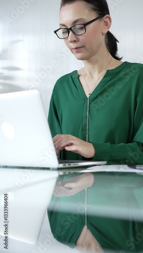 Businesswoman, wearing green blouse, is typing on laptop keyboard, preparing taxes and audit reports, with documents on glass office desk, vertical portrait view. Taxes, audit in business