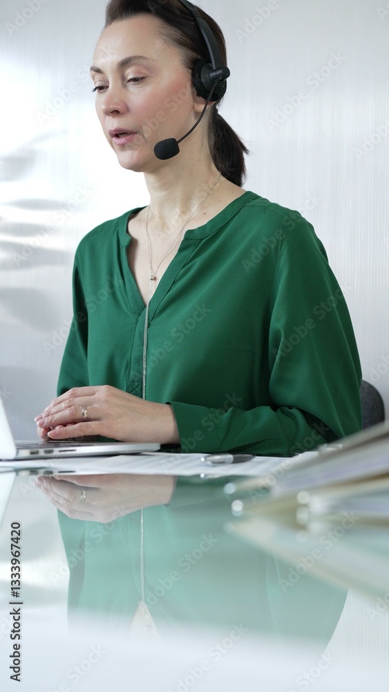 Businesswoman in green blouse is wearing a headset, focused on her laptop while efficiently working with laptop in a modern office environment, vertical portrait. Business people, call center concept