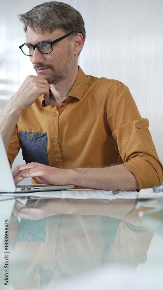 Focused businessman in yellow casual shirt is using laptop and thinking with hand on chin, working on a project in a modern office, vertical portrait. Business people concept